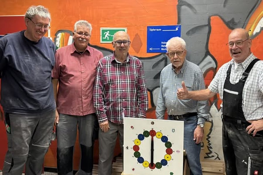 Fünf Männer stehen vor einer orangefarbenen Wand mit Graffiti und einem grünen Notausgangsschild, vor ihnen eine Scheibe mit bunten Kugeln auf einem Ständer.