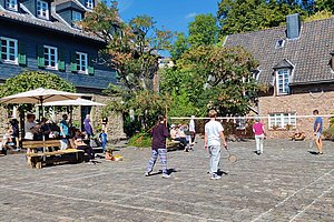 Mehrere Personen spielen Badminton auf einem gepflasterten Hof vor Fachwerkhäusern unter blauem Himmel.