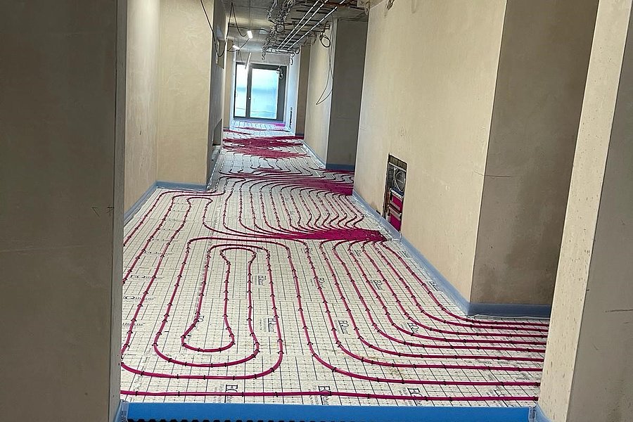 Flur mit sichtbaren roten Heizungsrohren auf dem Boden, die in Schleifen verlegt sind, weiße Wände und Decke mit sichtbaren Kabeln.