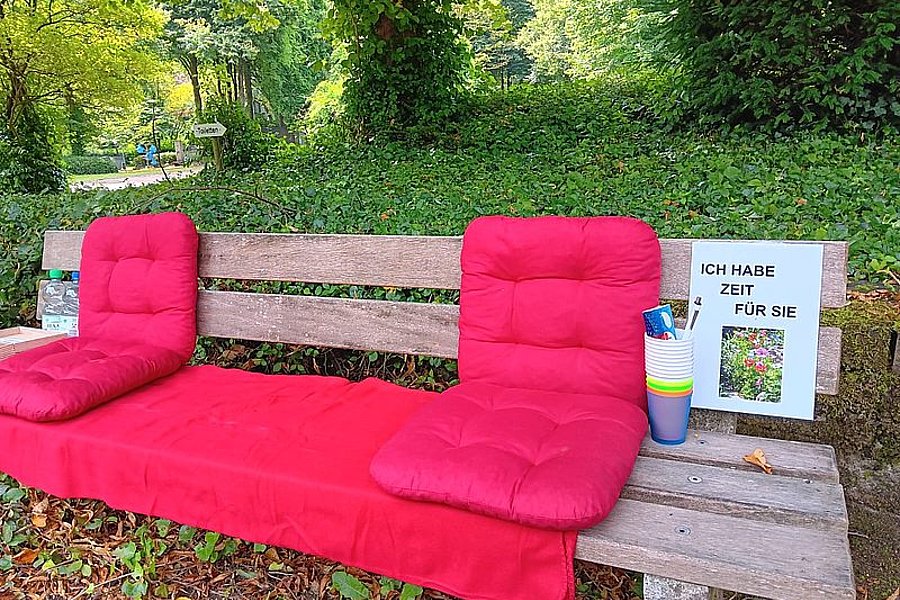 Holzbank im Park mit roten Sitzkissen und einem Schild mit der Aufschrift 'Ich habe Zeit für Sie' neben bunten Bechern