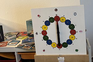 Bunte Spielscheibe mit Zahlen von 1 bis 16, zwei schwarzen Zeigern auf 12 Uhr, umgeben von kleinen Tier- und Glückssymbolen, auf einem weißen Ständer vor Bücherregal.