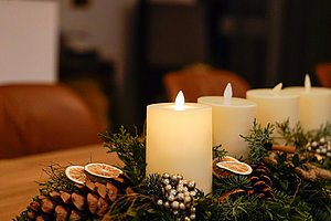 Vier brennende Kerzen stehen auf einem Adventsgesteck mit Tannenzweigen, Zapfen und getrockneten Orangenscheiben auf einem Holztisch.