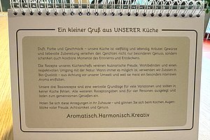 Aufgeschlagenes, spiralförmig gebundenes Kochbuch mit Text über die Vielfalt und Qualität der Küche sowie dem Motto 'Aromatisch.Harmonisch.Kreativ'