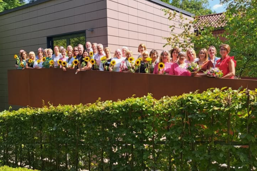 Gruppe von Menschen steht auf einer Terrasse vor modernem Gebäude, jede Person hält eine Sonnenblume oder einen Blumenstrauß.