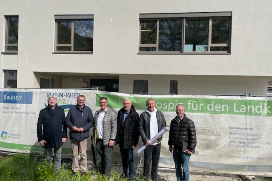 Sechs Männer in Jacken stehen vor einem Bauzaun mit Banner vor einem modernen Gebäude mit großen Fenstern.