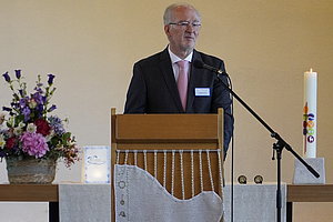 Mann in dunklem Anzug und Krawatte steht hinter einem Rednerpult mit weißem Tuch, daneben ein Mikrofon und eine Kerze, im Hintergrund bunte Glasfenster.