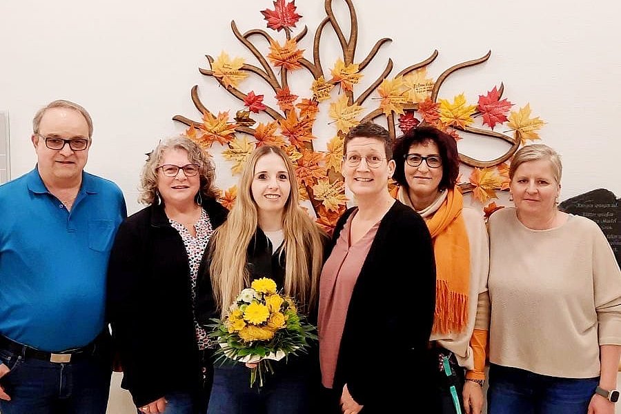 Sechs Personen stehen vor einer Wand mit einer dekorativen Metallskulptur aus herbstlichen Blättern. Eine Frau in der Mitte hält einen Blumenstrauß mit gelben Blumen.