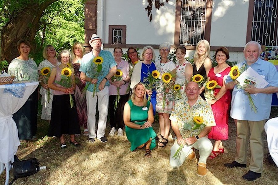 Gruppe von Menschen vor einem Haus, die jeweils eine Sonnenblume halten, zwei Personen knien vorne auf dem Gras