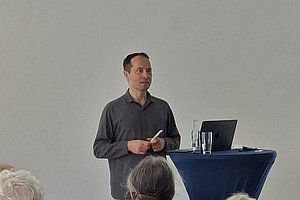 Mann steht vor weißer Wand hinter Stehtisch mit Laptop und Glas und hält Stift in der Hand