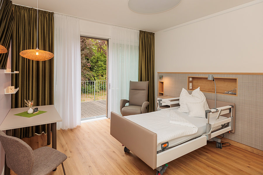 Krankenhauszimmer mit Pflegebett, Sessel, Schreibtisch, Stuhl, Vorhängen und Blick auf Garten durch offene Tür.