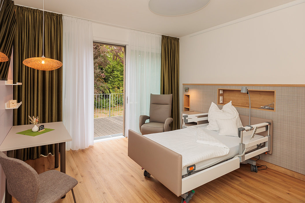 Krankenhauszimmer mit Pflegebett, Sessel, Schreibtisch, Stuhl, Vorhängen und Blick auf Garten durch offene Tür.