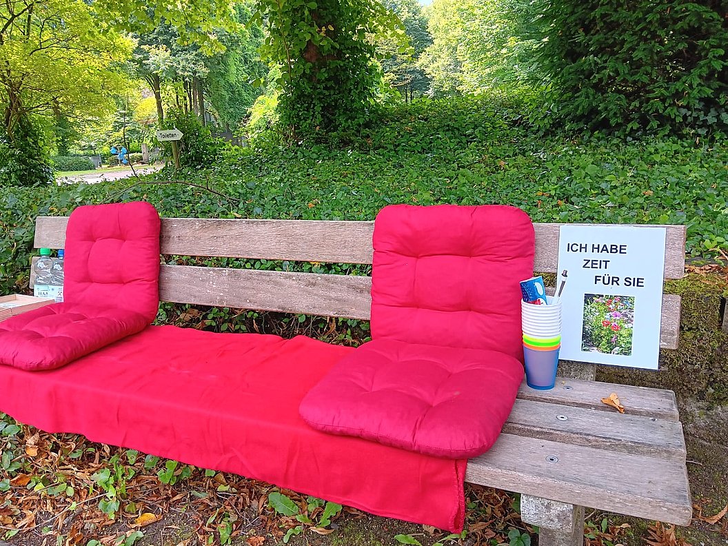Holzbank mit roten Sitz- und Rückenpolstern, daneben ein Schild mit der Aufschrift 'ICH HABE ZEIT FÜR SIE' und bunte Becher mit Stiften