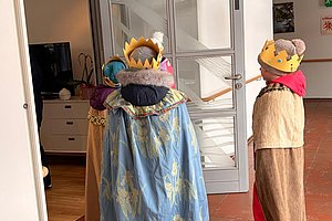 Drei Kinder mit Kronen und Umhängen stehen in einem Flur vor einer Glastür, zwei von ihnen blicken in einen Raum mit Fernseher und Kommode.