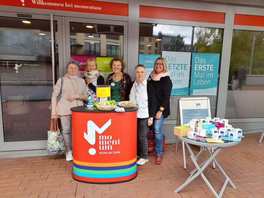 Fünf Personen stehen hinter einem roten Infostand mit der Aufschrift 'momentum Kirche am Osttor' vor einem Gebäude mit Glasfront und Werbeplakaten.