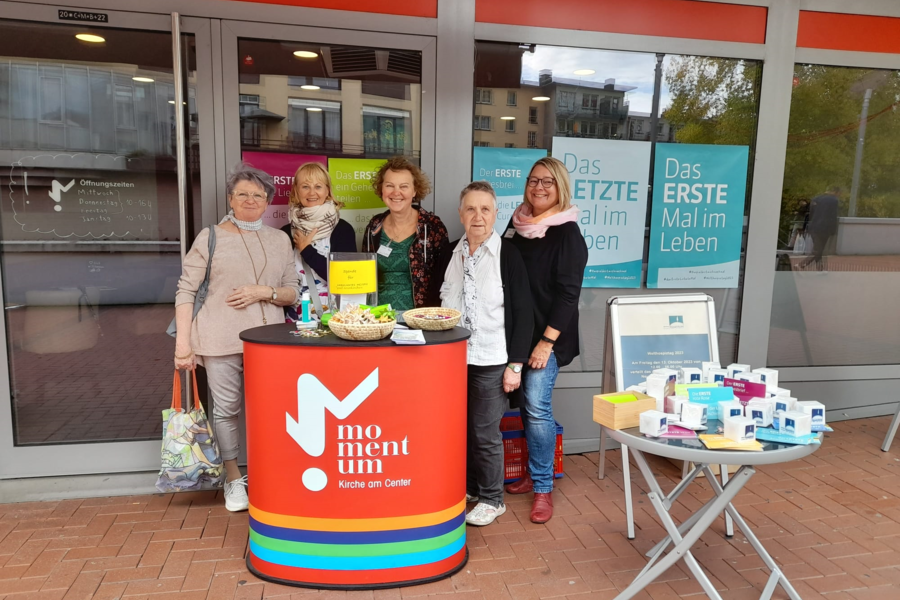 Fünf Personen stehen hinter einem roten Infostand mit der Aufschrift 'momentum Kirche am Osttor' vor einem Gebäude mit Glasfront und Werbeplakaten.