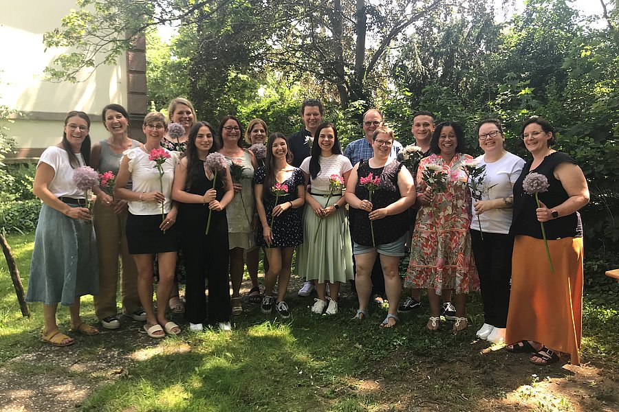 Gruppe von Menschen steht im Garten, jede Person hält einen Blumenstrauß, im Hintergrund Bäume und ein Haus.