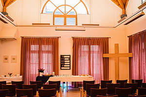 Innenraum einer Kapelle mit Holzbalkendecke, Stuhlreihen, Altar und großem Holzkreuz an der rechten Wand