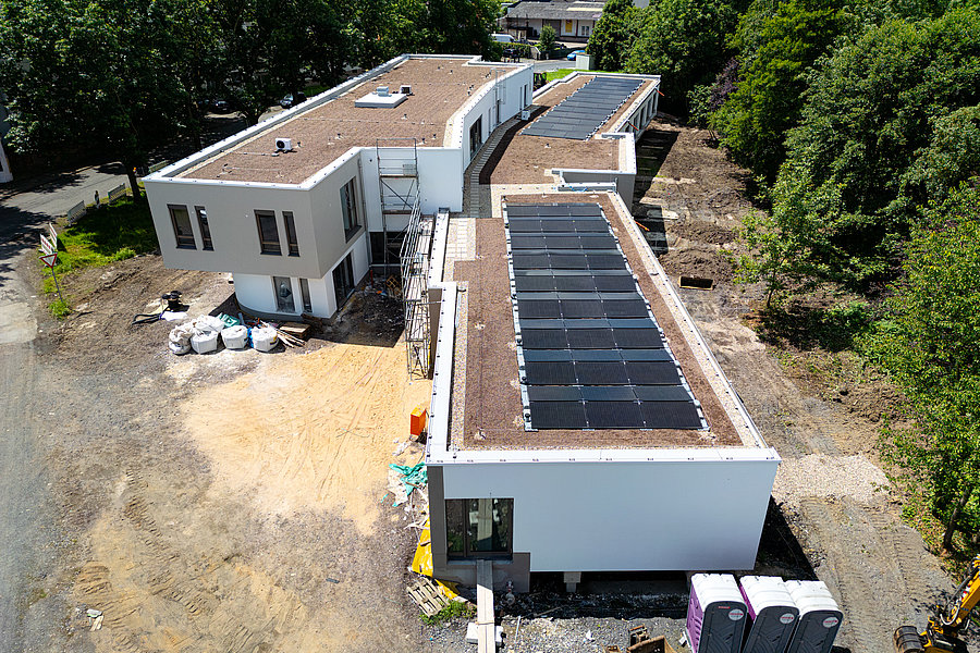 Luftaufnahme von zwei modernen, weißen Flachdachgebäuden, eines mit Solarpaneelen auf dem Dach, umgeben von Bäumen und Baustellenmaterial.
