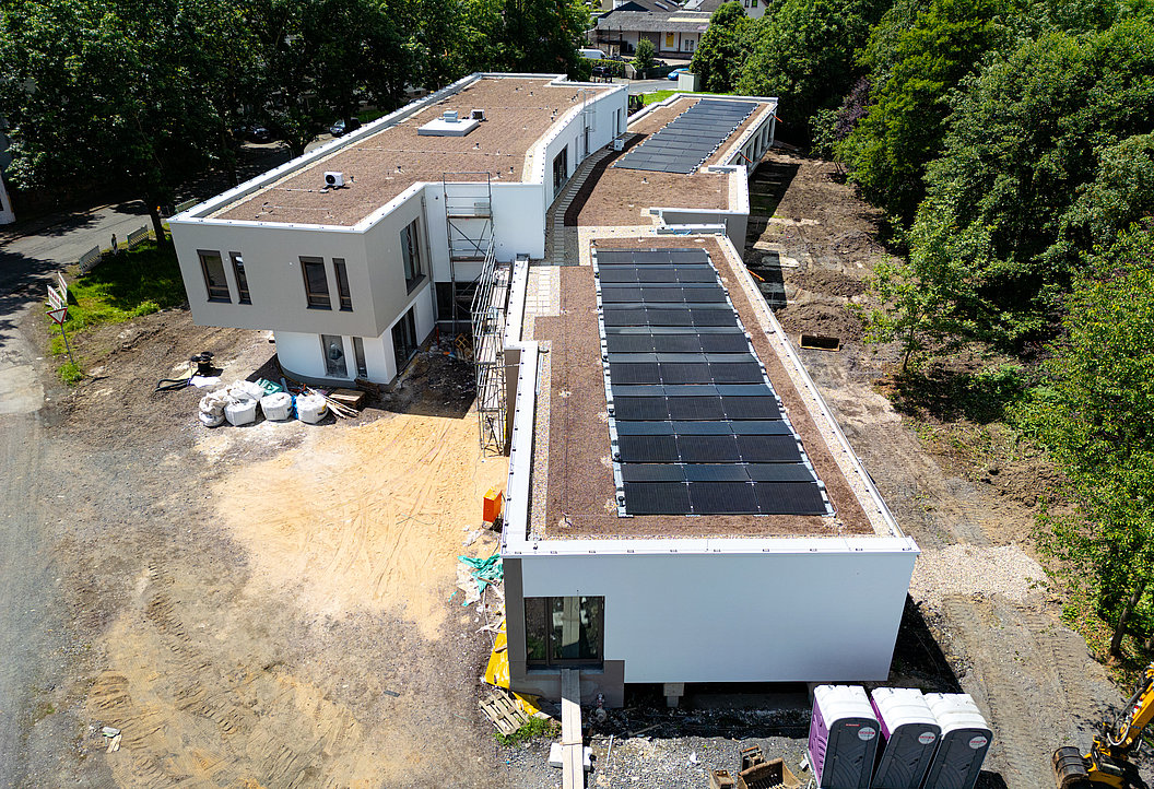 Luftaufnahme von zwei modernen, weißen Flachdachgebäuden, eines mit Solarpaneelen auf dem Dach, umgeben von Bäumen und Baustellenmaterial.