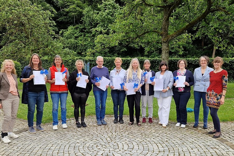Gruppe von elf Personen steht auf gepflastertem Platz vor Bäumen, einige halten Papiere in der Hand.