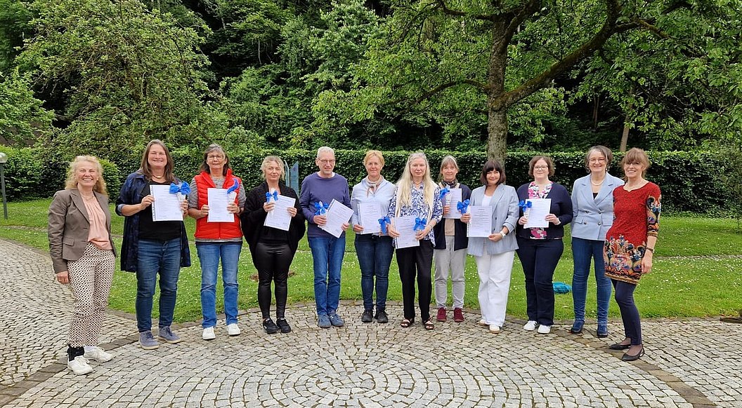 Gruppe von elf Personen steht auf gepflastertem Platz vor Bäumen, einige halten Papiere in der Hand.