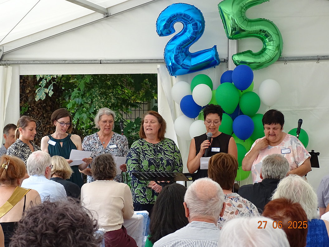 Gruppe von Menschen sitzt und steht in einem Zelt vor Publikum mit blauen und grünen Luftballons in Form der Zahl 25