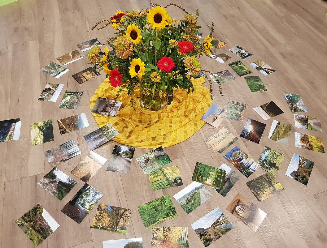 Strauß aus Sonnenblumen und roten Blumen auf gelbem Tuch, umgeben von mehreren Fotos auf Holzboden.