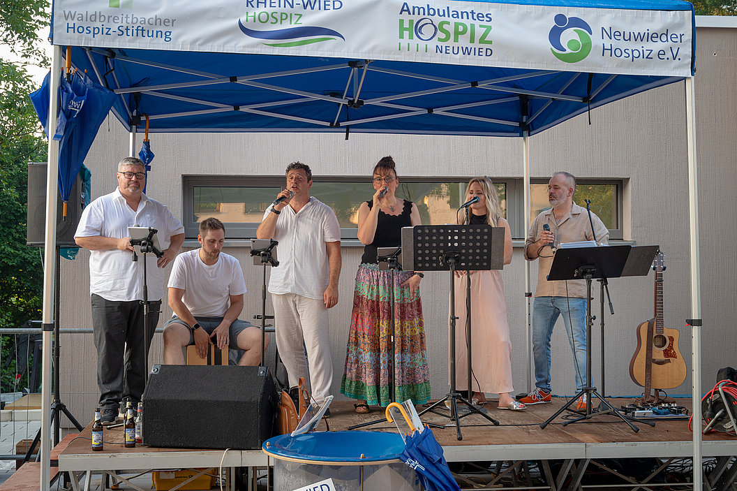 Sechs Personen auf einer Bühne unter einem blauen Zelt mit Logos von Hospiz-Organisationen, einige singen oder sprechen in Mikrofone, eine Person spielt Cajón.
