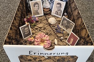 Schachtel mit Spitzenmuster, darin fünf gerahmte Porträtfotos, getrocknete Blumen, Muscheln und Steine, vorne Schriftzug 'Erinnerung'.