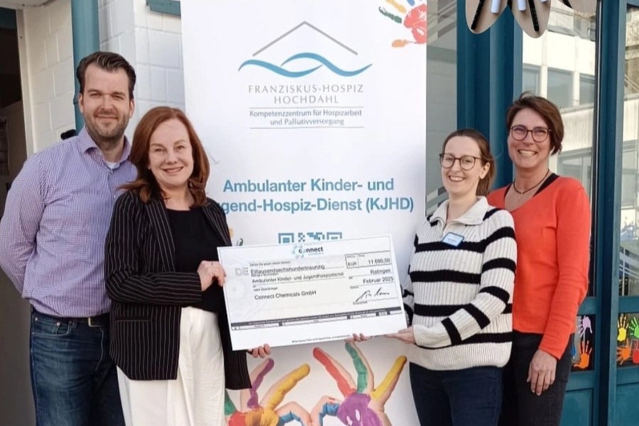Vier Personen stehen vor einem Banner des Kinder- und Jugend-Hospiz-Dienstes, zwei halten gemeinsam einen großen Scheck, bunte Hände formen ein Herz auf dem Banner.