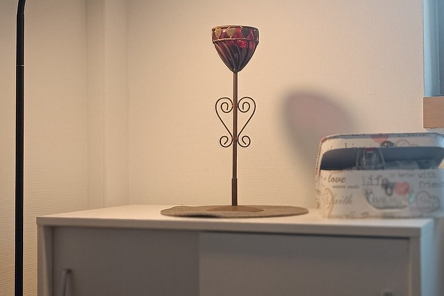 Kerzenhalter mit verziertem Stiel auf weißem Schrank, daneben weiße Box mit Schrift, links Stehlampe, Wand mit blau-weißem Wandbehang