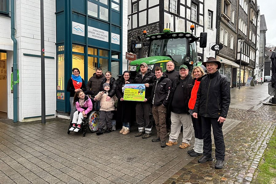 Gruppe von Menschen vor einem Fachwerkhaus und einem grünen Traktor, eine Person im Rollstuhl hält ein buntes Plakat.