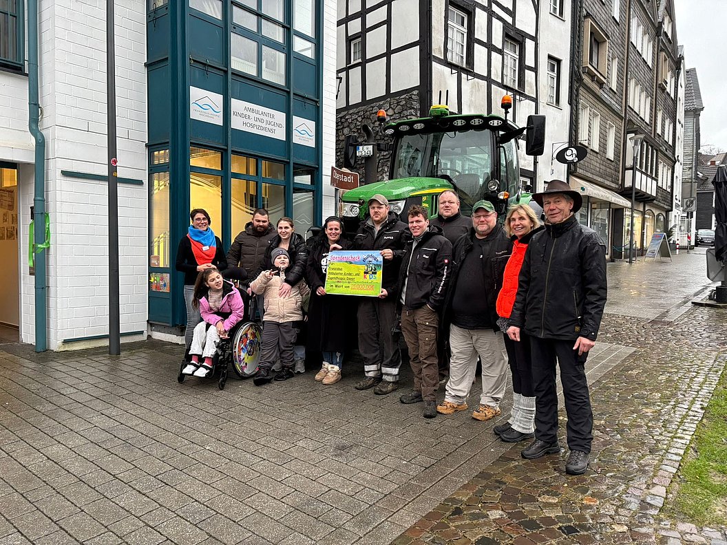 Gruppe von Menschen vor einem Fachwerkhaus und einem grünen Traktor, eine Person im Rollstuhl hält ein buntes Plakat.