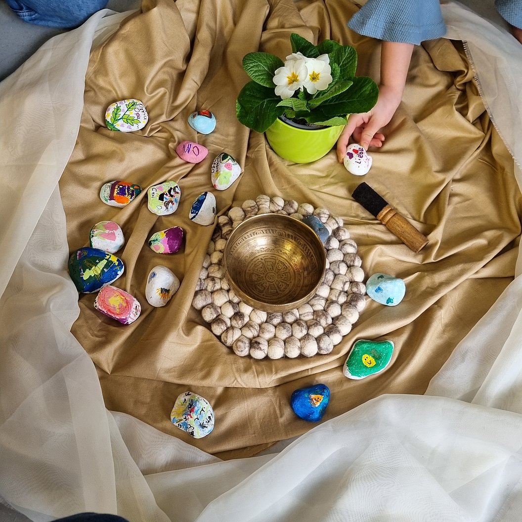 Goldfarbene Stofffläche mit einem runden Messingklangschalen-Set, umgeben von bemalten Steinen und einer grünen Topfpflanze mit weißen Blüten. Eine Hand legt einen bemalten Stein dazu.