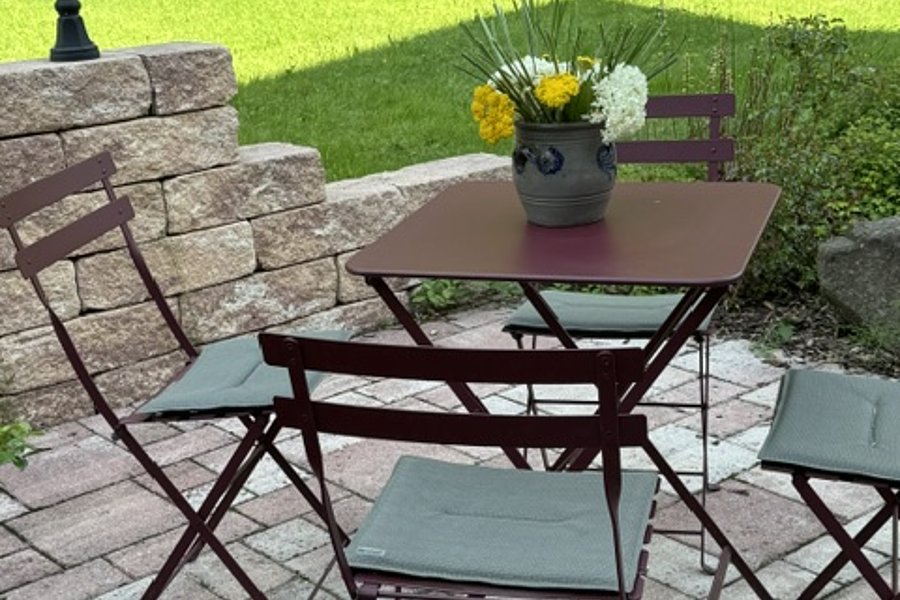 Garten mit gepflastertem Boden, einem runden Tisch mit Blumenvase und vier Stühlen mit Sitzkissen, umgeben von einer niedrigen Steinmauer und grünem Buschwerk
