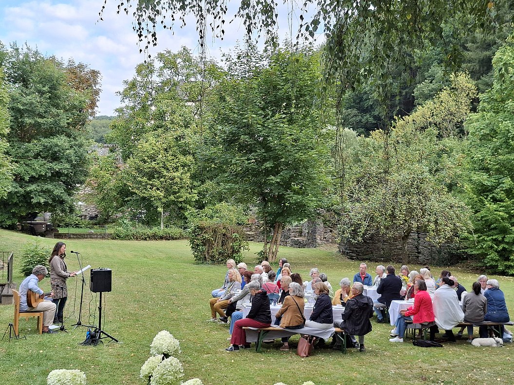 Mehrere Personen sitzen auf Bänken im Freien und hören einer Frau zu, die mit Mikrofon und Gitarre vor ihnen steht.