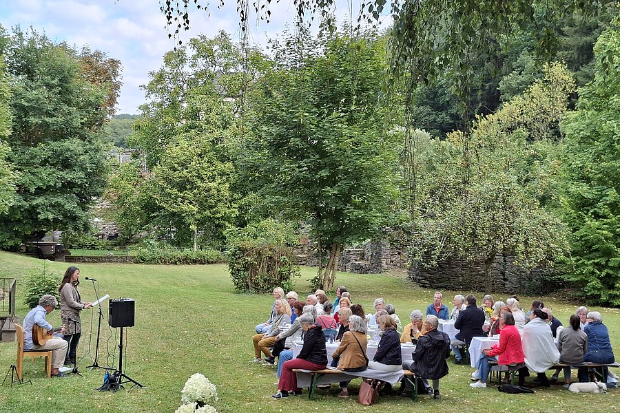 Mehrere Personen sitzen auf Bänken im Freien und hören einer Frau zu, die mit Mikrofon und Gitarre vor ihnen steht.