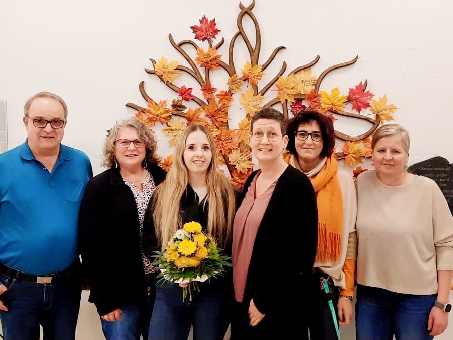 Sechs Personen stehen vor einer Wand mit einer dekorativen Metallskulptur aus herbstlichen Blättern. Eine Frau in der Mitte hält einen Blumenstrauß mit gelben Blumen.