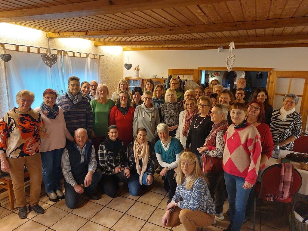 Gruppenfoto von 24 Personen in einem Raum mit Holzbalkendecke und Kachelfußboden, einige sitzen, andere stehen, Kleidung in verschiedenen Farben und Mustern.