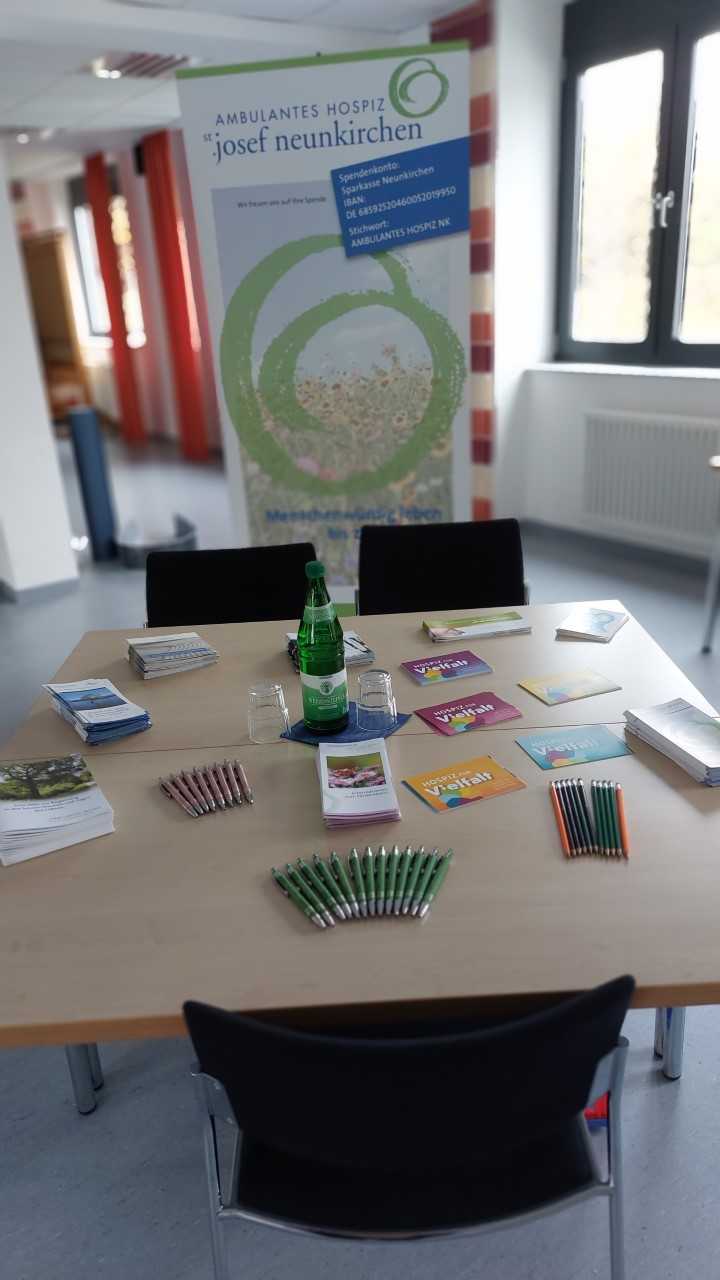 Tisch mit grünen Kugelschreibern, Flyern und einer Wasserflasche vor einem Banner des Ambulanten Hospiz Josef Neunkirchen in einem hellen Raum mit Fenstern.