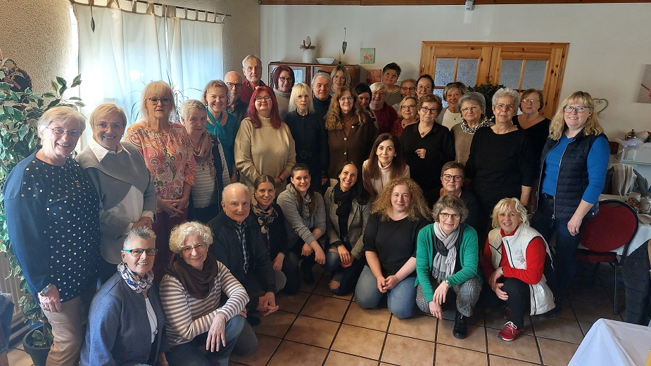 Gruppenfoto von 27 Personen in einem Raum mit Kacheln, Vorhängen und Holzmöbeln, einige sitzen, andere stehen, verschiedene Kleidung und Frisuren sichtbar.