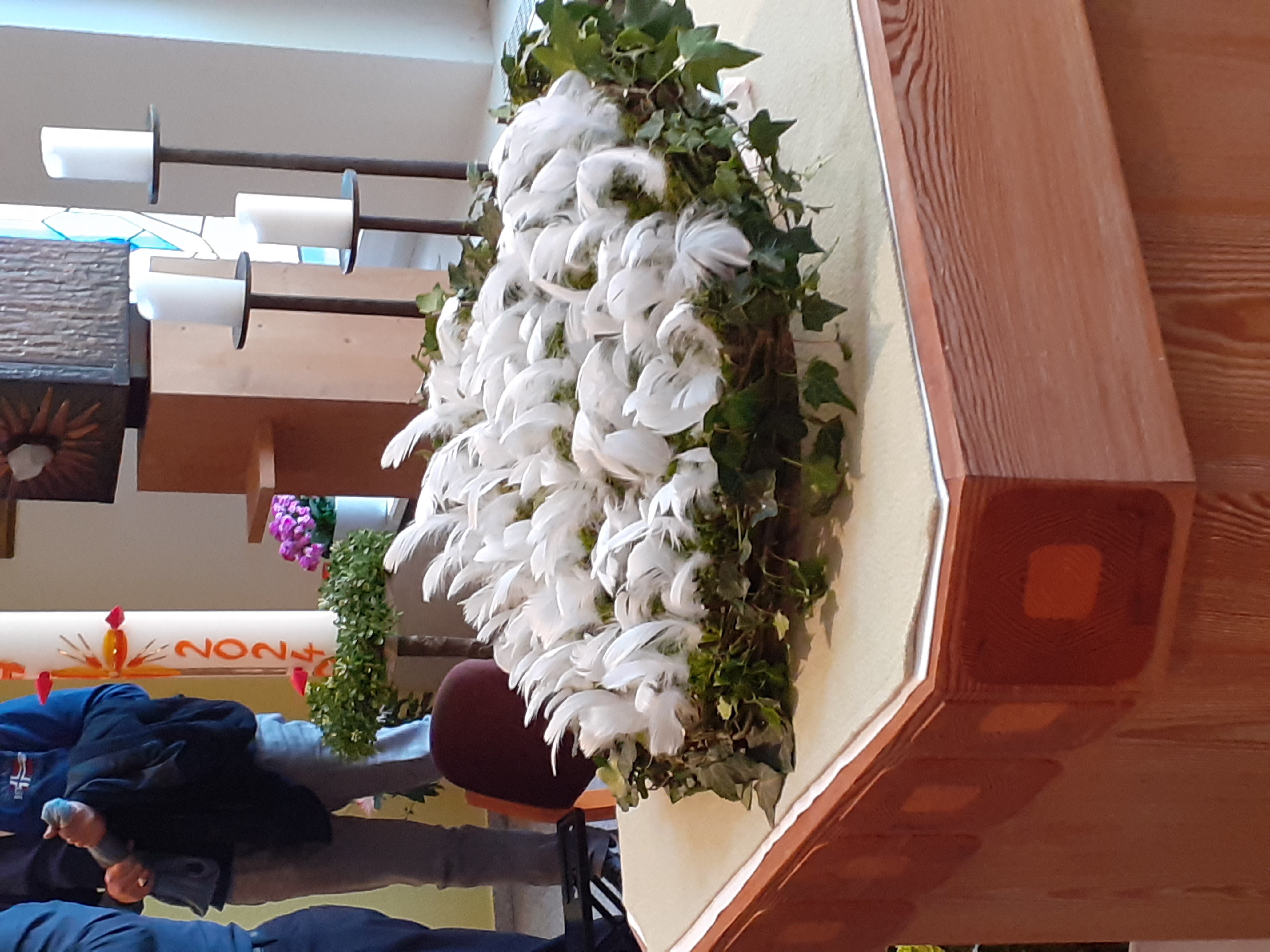 Kranz mit weißen Federn und grünen Blättern auf einem hölzernen Altar in einem Kirchenraum.