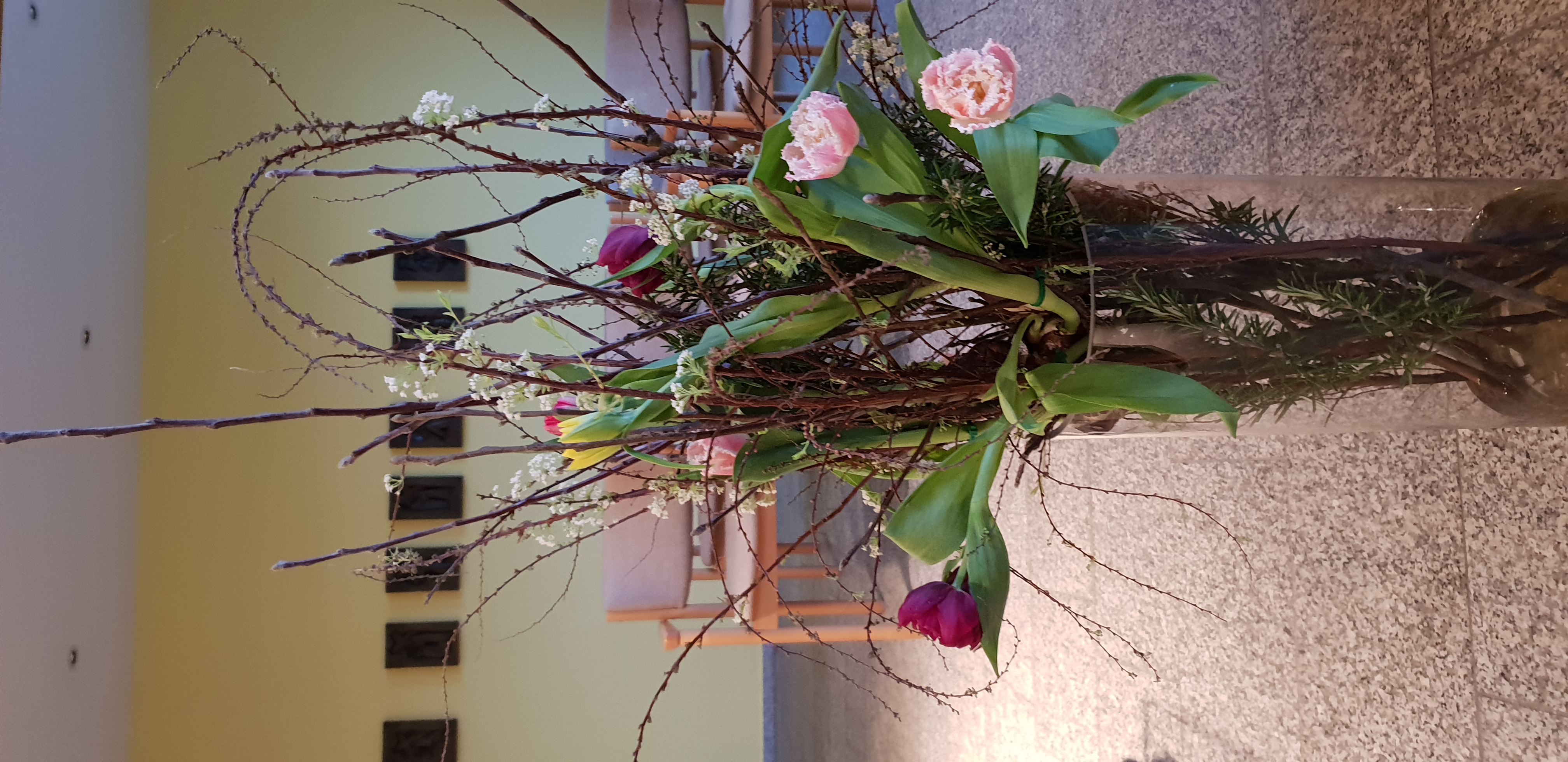 Blumengesteck mit Zweigen, grünen Blättern und rosa sowie roten Tulpen in einem hohen Glasvasenständer in einem Raum mit Stühlen und Bildern an der Wand
