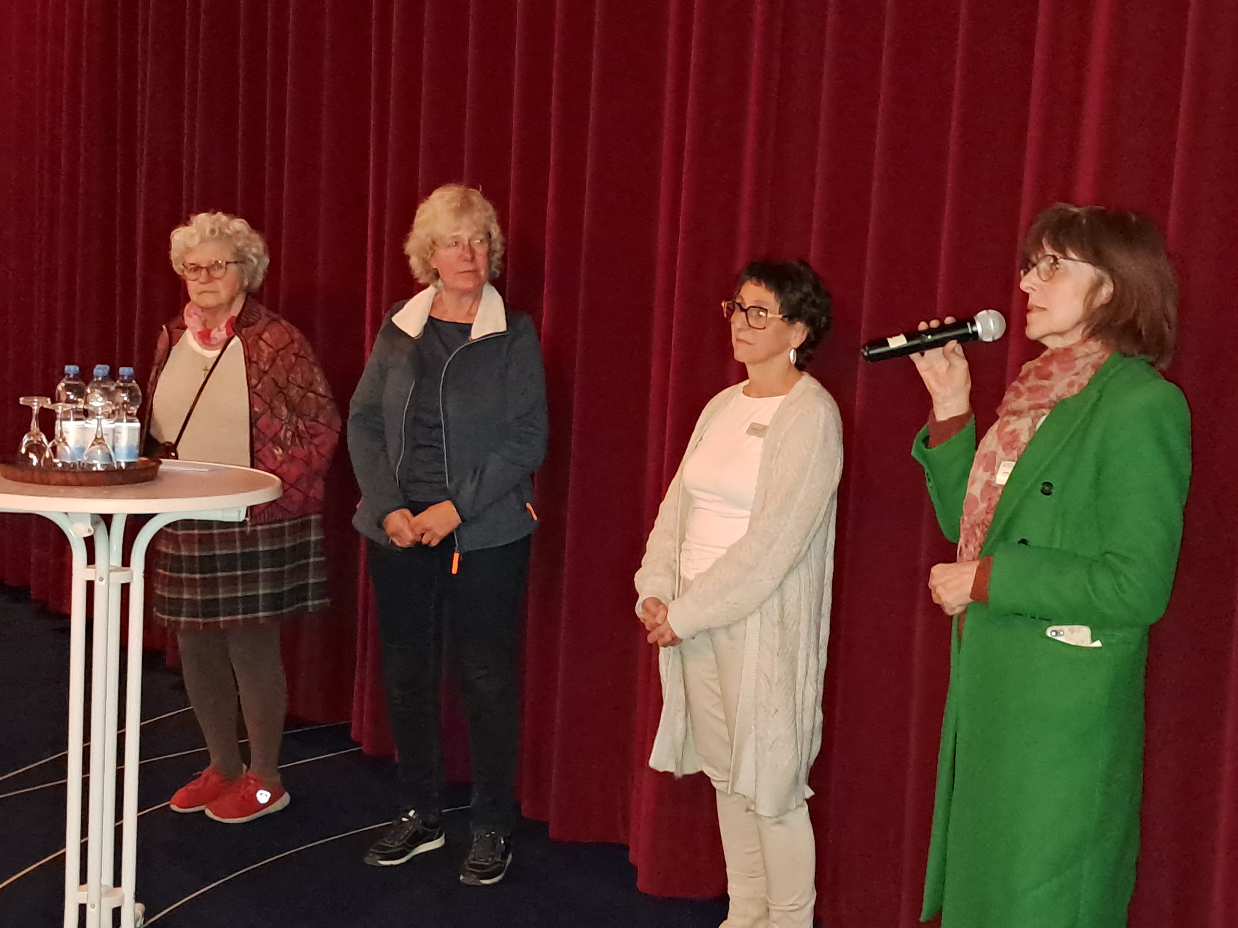 Vier Frauen stehen vor einem roten Vorhang, eine hält ein Mikrofon, auf einem Stehtisch stehen Wasserflaschen und Gläser.