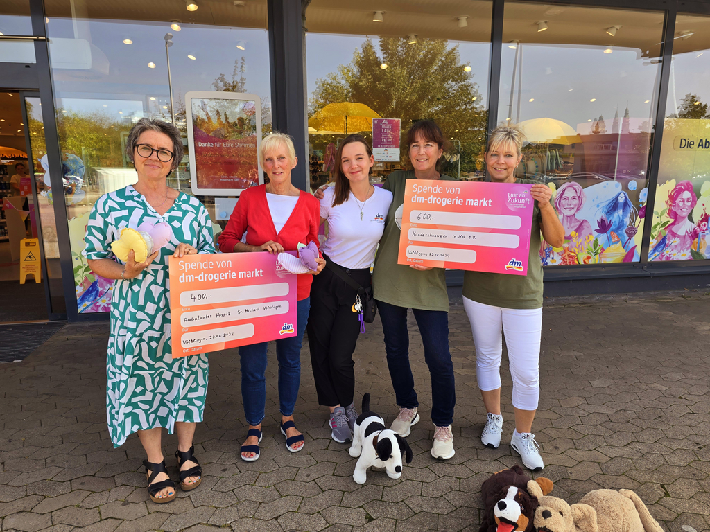 Fünf Personen stehen vor einem Geschäft, zwei halten große pinke Spendenschecks, eine Person hält ein Stofftier, ein kleines Stoffhundspielzeug liegt auf dem Boden.