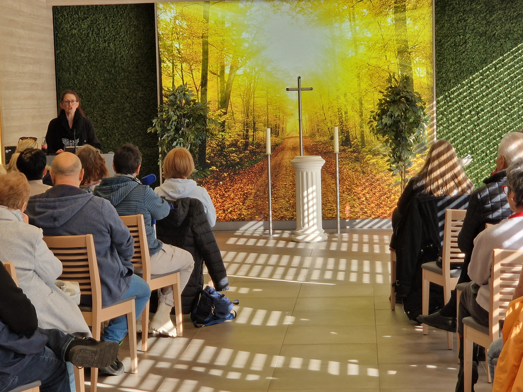 Mehrere Personen sitzen auf Holzstühlen in einem Raum mit grünem Pflanzenwand-Dekor und einem großen Wandbild eines herbstlichen Waldes mit Kreuz auf einem weißen Podest.