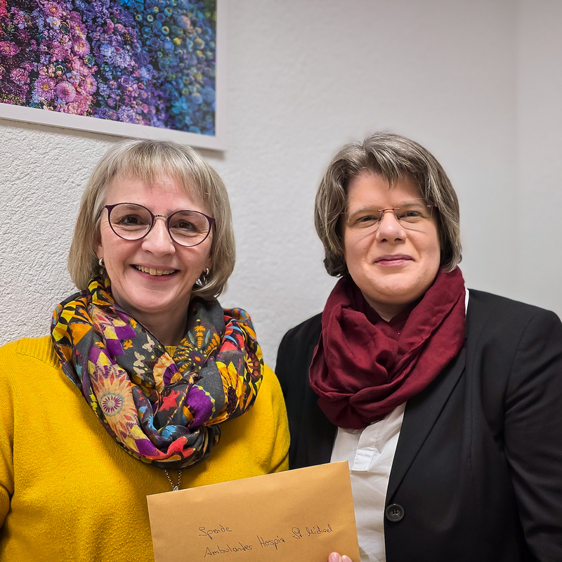 Zwei Frauen sitzen nebeneinander, eine trägt einen gelben Pullover und einen bunten Schal, die andere einen schwarzen Blazer und einen roten Schal. Eine hält einen braunen Umschlag mit handschriftlichem Text.