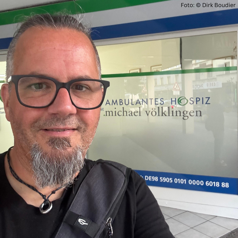 Oberkörper einer Person mit grauen Haaren und schwarzem T-Shirt vor einer Glaswand mit der Aufschrift 'AMBULANTES HOSPIZ'