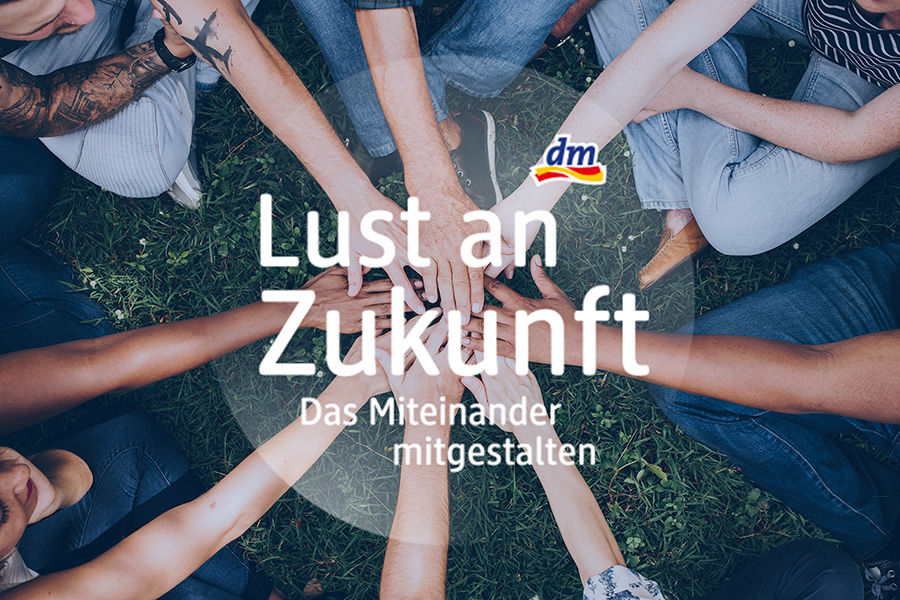 Mehrere Menschen sitzen im Kreis auf Gras und legen ihre Hände in die Mitte, darüber der Text 'Lust an Zukunft Das Miteinander mitgestalten' und das dm-Logo.