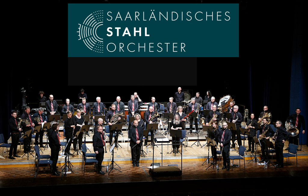Orchester auf Bühne mit verschiedenen Blasinstrumenten und Streichinstrumenten, im Hintergrund Banner mit Schrift 'Saarländisches Stahl Orchester'.